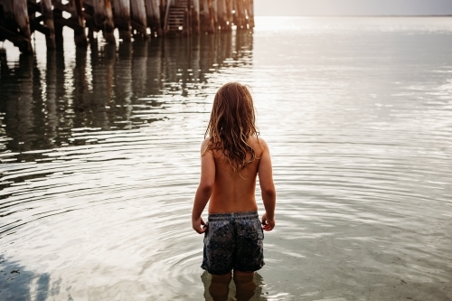 Young boy in shallow water looking out into ocean - Australian Stock Image