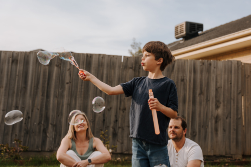 Young boy playing with bubbles in their yard while parents sit on the ground - Australian Stock Image