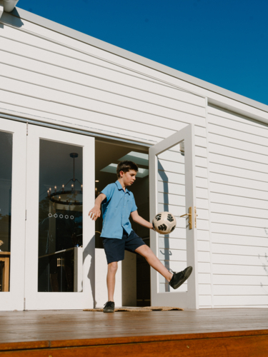 Young boy practicing soccer on sunlit deck outside home - Australian Stock Image