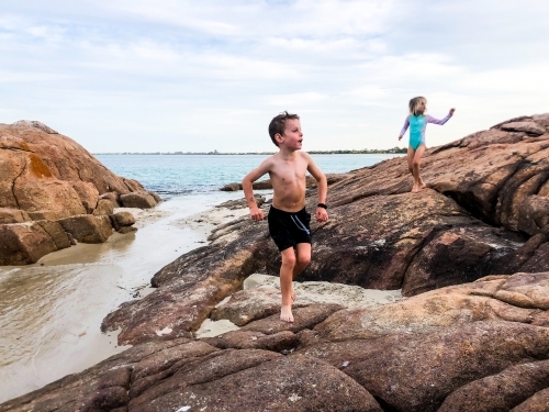 Young boy running over coastal rocks with young girl following him