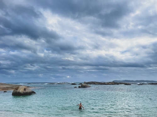 Young boy standing in ocean on overcast day with submerged granite rocks - Australian Stock Image