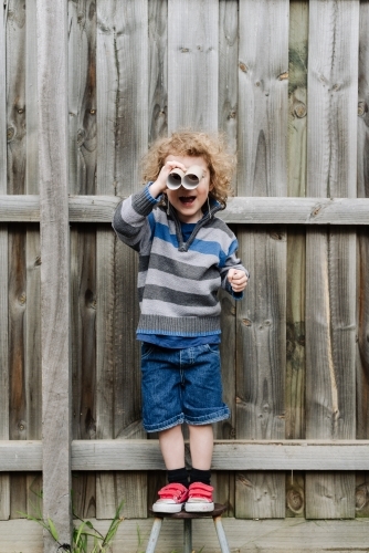 Young boy standing on a stool playing with cardboard toilet roll binoculars in the backyard - Australian Stock Image