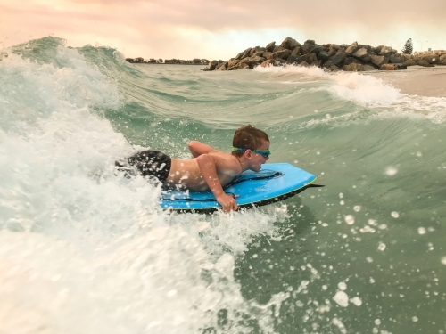Young boy surfing boogie board on beach wave with groin in background with hazy pastel sunset - Australian Stock Image
