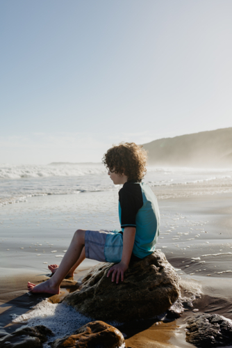 Young boy with curly hair sitting on a rock relaxing at the beach in the afternoon - Australian Stock Image