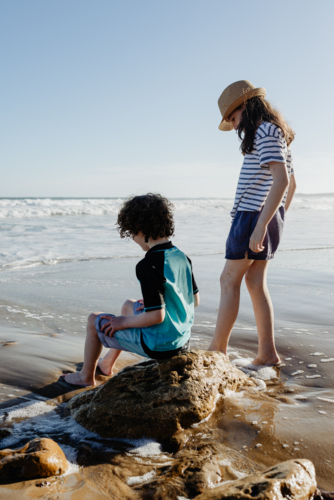 Young brother sitting on a rock and his sister (young boy and girl) together at the beach - Australian Stock Image