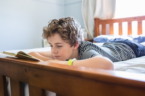 Young child lying on stomach on bed reading book natural light - Australian Stock Image