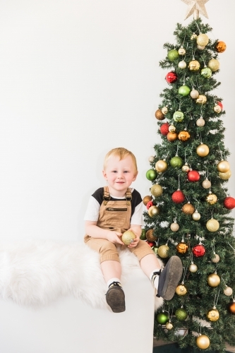 Young child sitting on rug holding bauble next to Christmas tree - Australian Stock Image