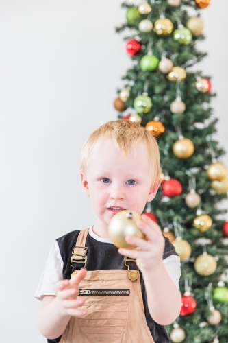 Young child standing in front of Christmas tree holding decoration - Australian Stock Image