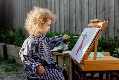 Young child wearing an art smock painting with an easel in the backyard garden at home - Australian Stock Image