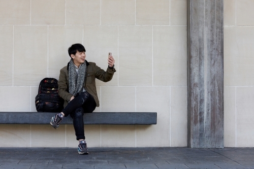 Young Chinese man sitting on bench using mobile phone - Australian Stock Image