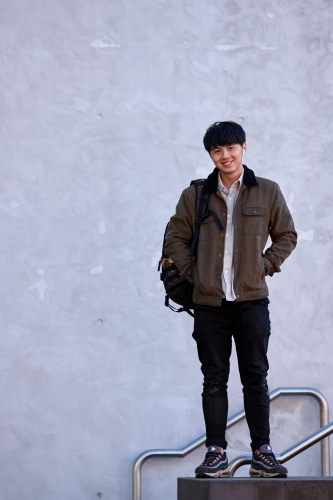 Young Chinese standing against concrete wall with backpack - Australian Stock Image