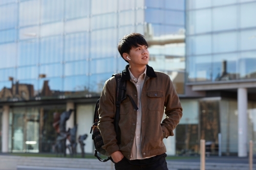 Young Chinese student standing with hands in pocket with listening to wireless headphones - Australian Stock Image