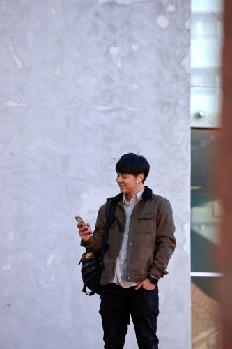 Young Chinese university student standing using mobile phone and wireless headphones - Australian Stock Image