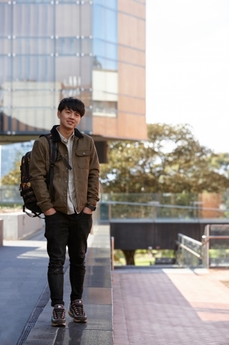 Young Chinese university student standing with hands in pocket listening to wireless headphones - Australian Stock Image