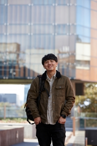 Young Chinese university student standing with hands in pocket listening to wireless headphones - Australian Stock Image