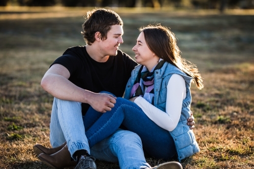 Young couple sitting close together on grass legs entwined smiling at each other - Australian Stock Image