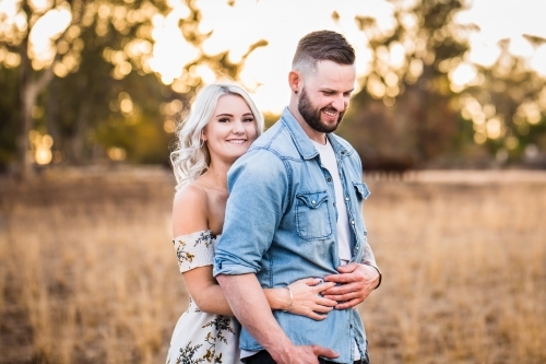 Young couple standing together with woman's arms around man's waist smiling - Australian Stock Image