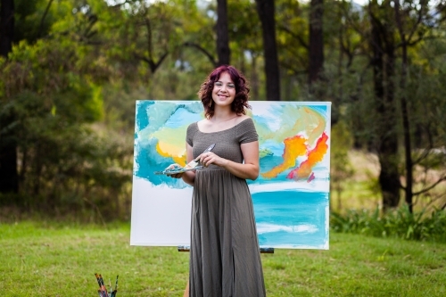 Young creative artist with curly purple hair working on canvas with acrylic paints - Australian Stock Image