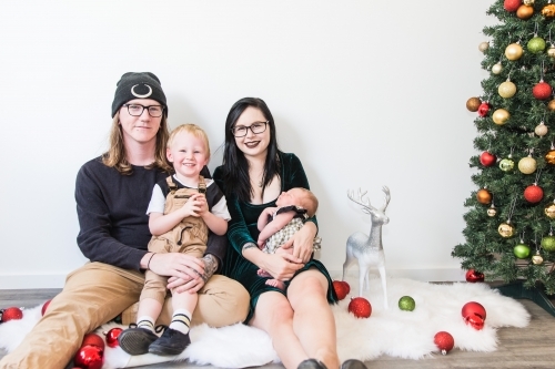 Young family sitting close together on rug at Christmas with tree and decorations - Australian Stock Image