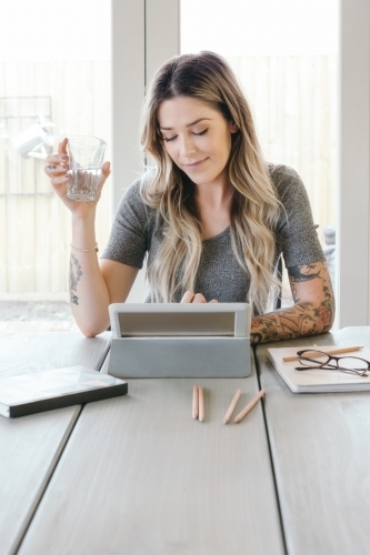 Young female working in a light bright space on a tablet with water in hand - Australian Stock Image