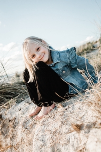 Young girl at the beach sitting on a sand dune looking at the camera wearing a denim jacket - Australian Stock Image