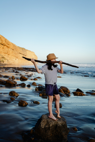 Young girl having fun playing at the beach standing on a rock holding a branch like a spear - Australian Stock Image