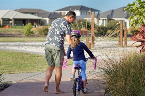 Young girl learning to ride her bicycle in the park with her dad helping - Australian Stock Image