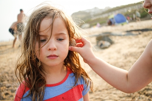 Young girl listening intently as older girl holds a shell to her ear at the beach - Australian Stock Image