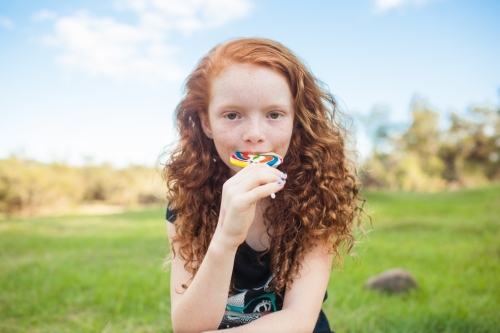 Young girl outside eating a rainbow lollipop - Australian Stock Image