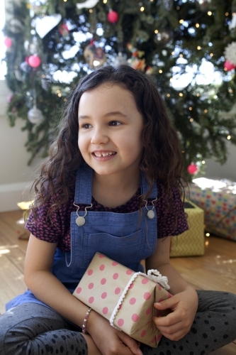 Young girl sitting in front of Christmas tree holding a present in her lap - Australian Stock Image