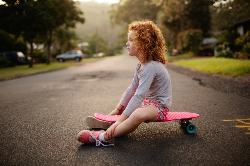 Young girl sitting on a skateboard on a road - Australian Stock Image