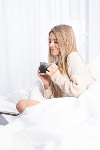 Young lady drinking hot drink in white bed - Australian Stock Image