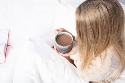 Young lady drinking hot drink in white bed from an overhead perspective - Australian Stock Image