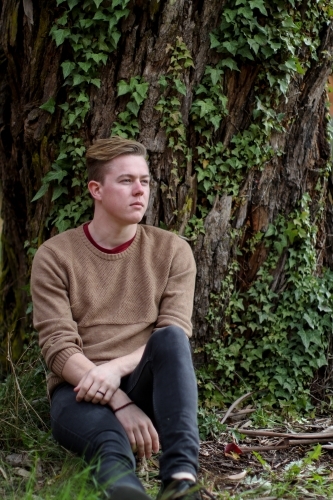 Young man sitting outdoors at the base of a tree contemplating life - Australian Stock Image