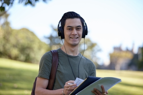 Young man with dark hair outside holding device wearing headphones at park - Australian Stock Image