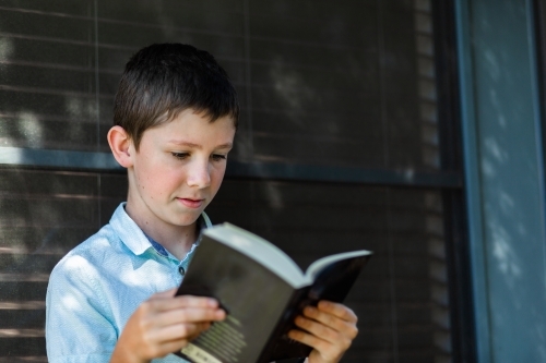 Young primary aged boy reading fiction book leaning against window outside home - Australian Stock Image