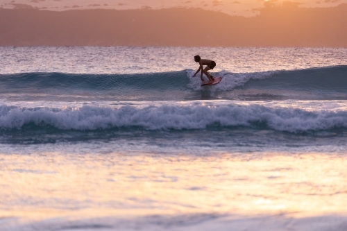 young surfer backlit surfing small waves at sunset