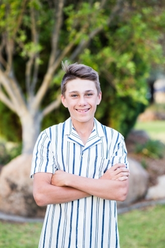 Young teenage boy standing with arms crossed smiling in garden - Australian Stock Image