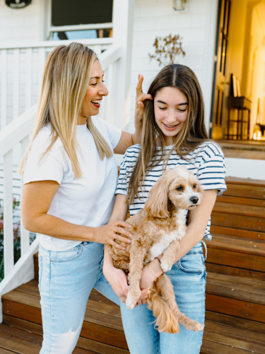 Young teenage girl carrying a dog while standing beside her mum outside the house. - Australian Stock Image