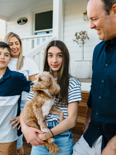 Young teenage girl carrying a dog while standing beside her parents and brother outside the house. - Australian Stock Image