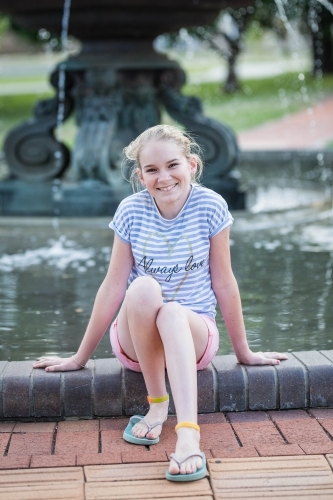 Young teenage girl sitting on edge of water fountain smiling - Australian Stock Image