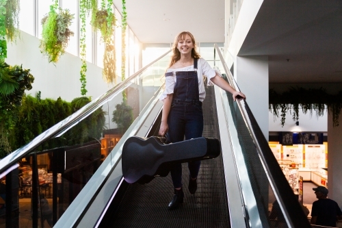 Young woman carrying guitar case down escalator in shopping center - Australian Stock Image