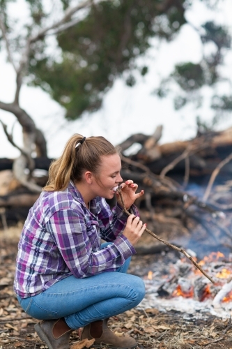 Young woman in checked shirt and jeans toasting marshmallows on campfire - Australian Stock Image