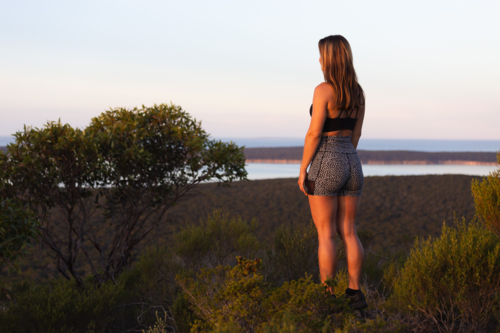 Young woman in fitness wear looking at scenic view over the sea from a mountain lookout at sunset - Australian Stock Image