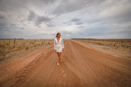 Young woman in white walking barefoot on rural dirt road - Australian Stock Image