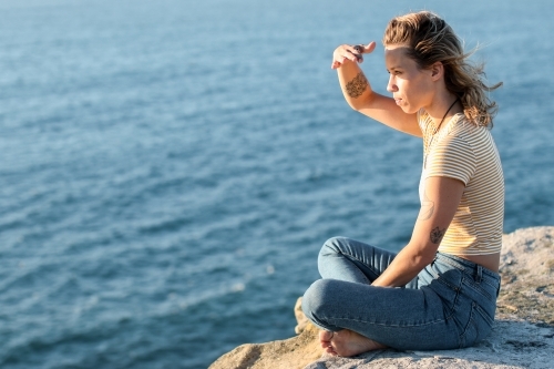 Young woman sitting on coastal clifftop at sunrise looking out to sea - Australian Stock Image