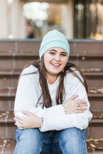 Young woman sitting on steps leaning forward wearing beanie smiling - Australian Stock Image