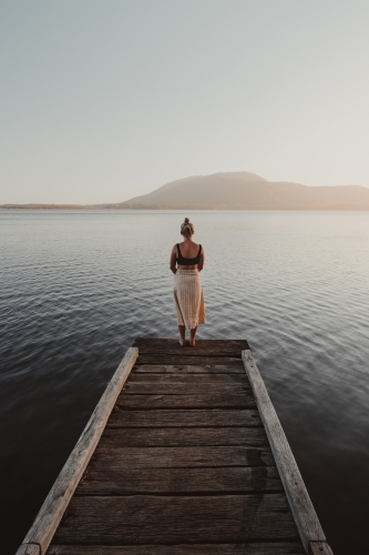 Young woman standing at the end of a jetty looking over a lake with a mountain in the background. - Australian Stock Image