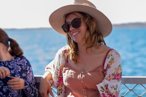 young woman wearing hat and sunglasses sitting with water in background - Australian Stock Image