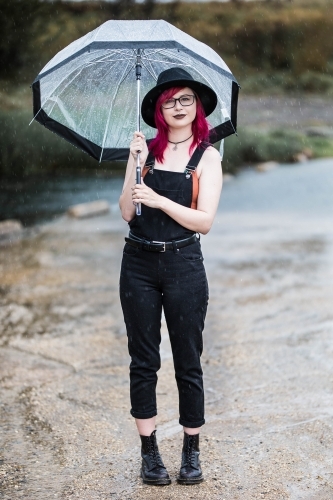 Young woman with pink hair standing near water holding umbrella in rain - Australian Stock Image